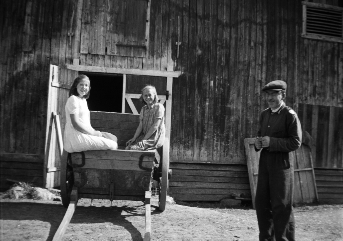 Ingrid Olstad og Mildrid Granberg sitter i stuttkjerra utenfor låven på Søndre -Tomter (Ner-Tomter) ca 1950. Odd Olstad til høyre. 