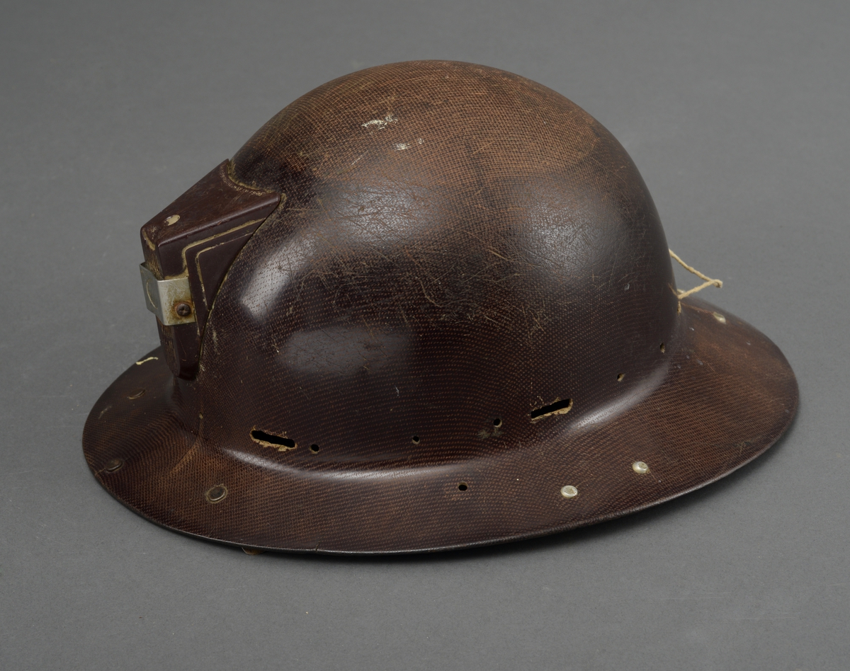 Gruvehjelm med påsatt del avplast og jern for feste av skjerm på undersiden av bremmen. Lampefeste på front- og ledningsfeste bak på hjelmen.