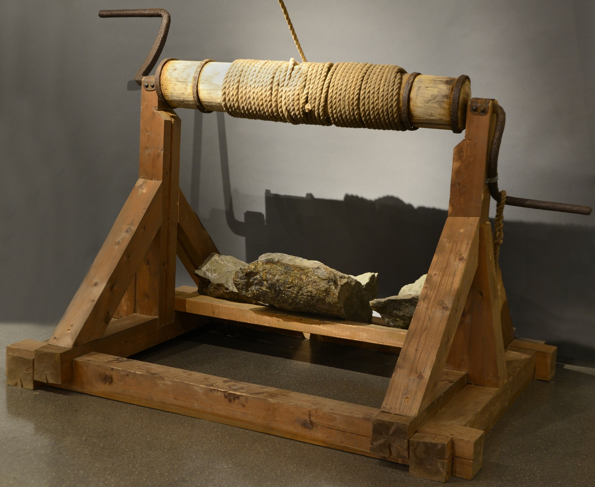 Heiseanretning av tre med sveiver i jern. Tauverk er festet til en vinsj som er montert i taket i utstillingen.