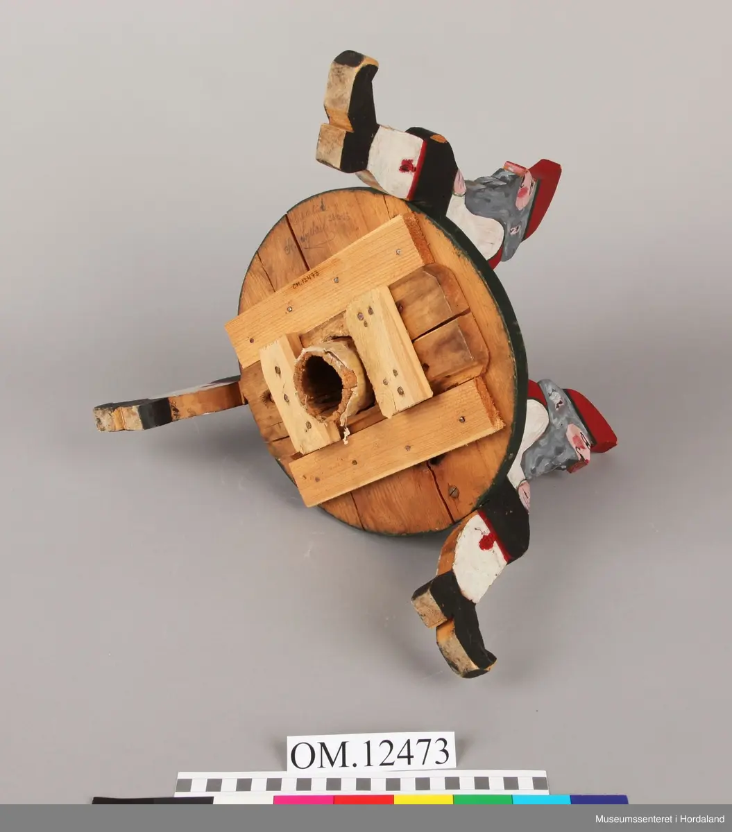 3 nisser i utskårne i tre som sit på ei rund plate. Nissaane og plata er skårne ut i 2 cm tjukk plank. Nissane er malte i ulike fargar. Plata er malt svart. Brun tape rund holet til juletreet.