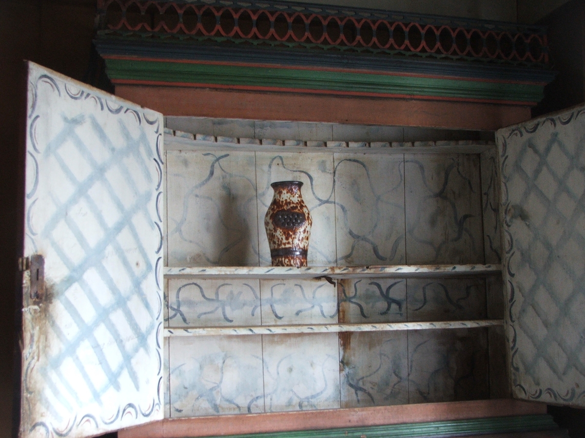 Todelt skap i tre. To skapdører øverst, 8 små skuffer i midten og fire større skuffer nederst. Oppheng til skjeer i øverste skap. Utskjæringer i tre øverst. Grønnmalte fyllinger dekorert med små blomster.