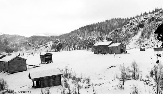 Stabbforsmoen gard i Hemnes statsskoger.  Fotografiet er tatt fra et høyt ståsted mot en snødekt voll med flere laftete bygninger.  I forgrunnen et lite enetasjes hus med overkraget gavl, bakenfor dette en låve og et loft.  Mer til høyre i bildet et stallåvekompleks med en hest utenfor, samt et par mindre bygninger.  I bakgrunnen en bergrygg med en del granskog. 