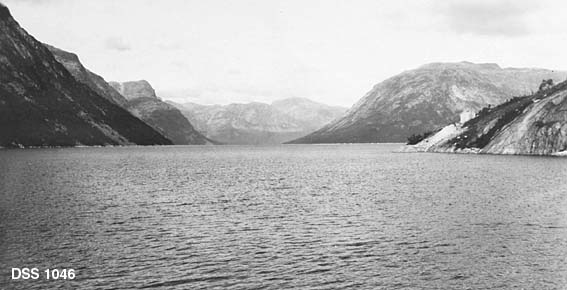 Fra Heggvassbotn i Heggmoen statsskog.  Fotografiet er et utsiktsbilde over en fjord, omgitt av forholdsvis bratte fjell med en del skog nederst mot vatnet.  På en bergnabbe til høyre i bildet ses gavlen på en liten stuebygning. 