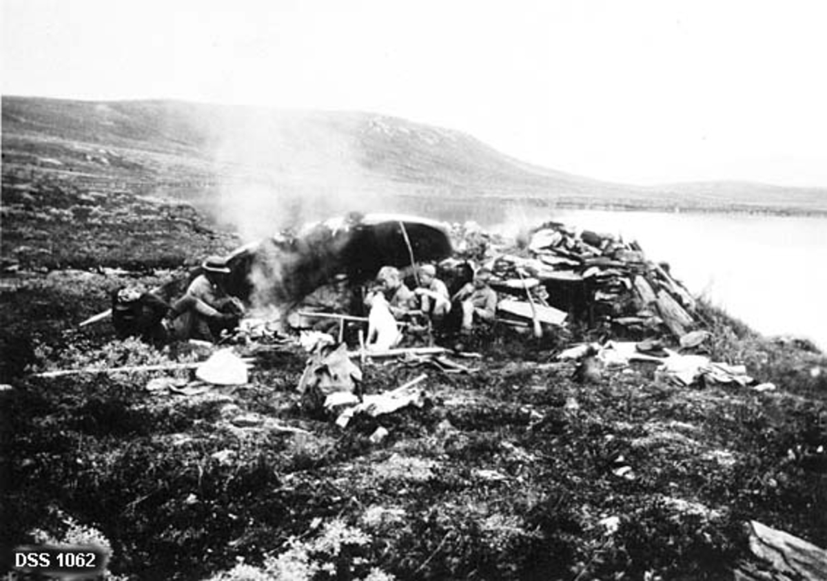 Rast ved Flattjonna i Hattfjelldal statsskog.  Fotografiet viser tre menn, tre gutter og en hund samlet om et måltid ved et bål.  Bålet er oppgjort i ly av en veltet båt, like inntil ei lav hytte, murt av naturstein.  I bakgrunnen Flattjønna omgit av lave bergrygger. 