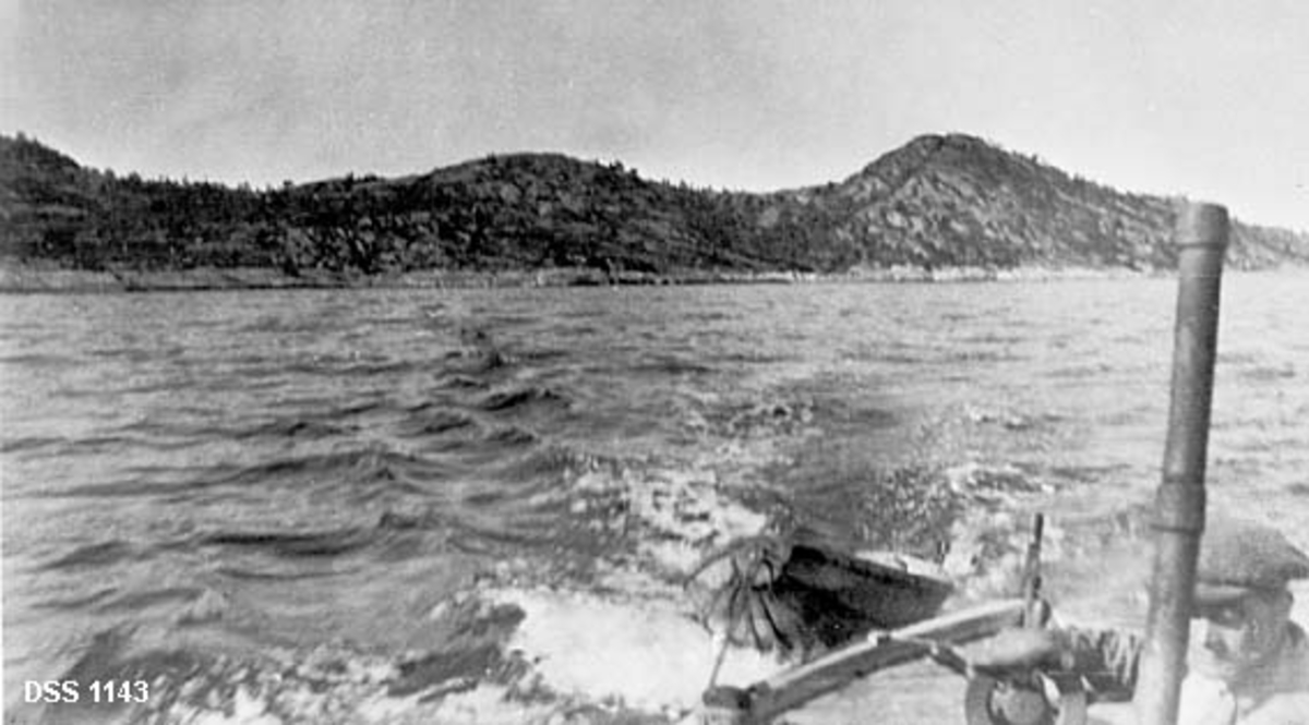 Fra Bekkenesholmen i Tysfjord.  Holmen har mange bergflater, men også en del furuskog. Fotografiet er tatt fra en motorbåt, der akterenden med eksosrør og styrmann, samt ei etterhengt jolle er synlige i forgrunnen. 