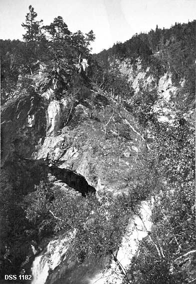 Landskapsfotografi fra Gråttådalen i Beiarn.  Fotografiet viser ei elv som renner i ei skråstilt revne nedover ei fjellside (øverst på bildet) for deretter å danne et fossefall rett nedover fjellsida (nederst på bildeflata).  Fossen er delvis dekt av bjørkebusker.  Det opprevne fjellet har også en del furutrær. 