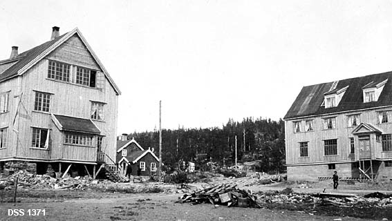 Administrasjonsbygningen og hotellet ved Grong gruvers anlegg i Gjersvika.  Fotografiet viser to store, nyoppførte og umalte trebygninger med vertikal ytterpanel.  Begge bygningene har to fulle etasjer, samt loft med arker som antakelig også har gitt lys til boligrom.  Bygningen til venstre later til å kvile på en kjeller av hoggen naturstein, mens den til høyre har betongmur og et vindfang som foreløpig mangler underbygning.  Mellom og litt bak bygningene skimtes et noe eldre trehus og ei telefonlinje som kommer til gards gjennom bjørkeskogen.  På gardsplassen er det mye bygningsavfall.  Det later til å gå en veg gjennom tunet, hvor en mann kommer gående. 