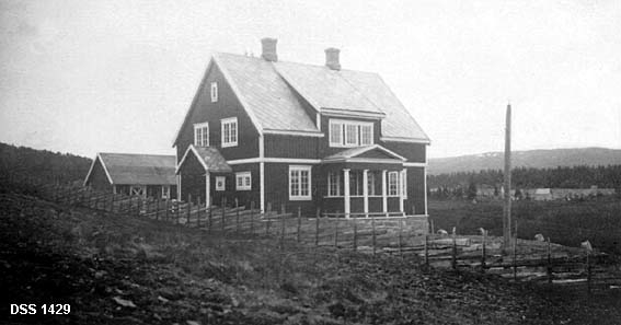 Skogvokterboligen i Snåsa. Halvannenetasjes villa i jordbrukslandskap. Bygningen er fotografert fra en nypløyd voll, og tomta avgrenses fra jordbruksarealet ved en skigard.  Bygningen er en trekonstruksjon med utvendig panel. Panelen er malt med en mørk farge, med unntak av hjørnebord, dør- og vindusomramming4er, vindskier og etasjeskiller, som er kvite.  På gavlenden er det oppført et vindfang foran ei inngangsdør, mens langsida mot hagen har en åpen veranda båret av fire kvitmalte trestolper.  Over verandaen er taket løftet litt for å gi plass til tre vinduer.  Taket er tekket med skiferheller. I bakgrunnen et enetasjes, bølgeblikktekket uthus. Til høyre skimtes også en bakkekam med åkrer og jordbruksbebyggelse, bakenfor dette skogkledde åser.