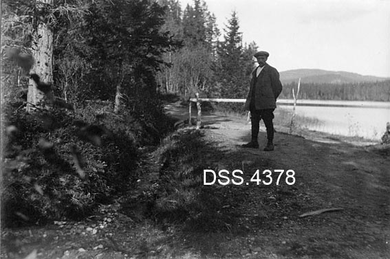 Skogsbilveg ved Ellingsvatnet i Hølonda i Sør-Trøndelag.  En mann med vindjakke og sixpencelue står på vegbanen.  Bak ham er vegen sperret på et sted der overflatevatn har gravd seg gjennom veglegemet.  Fotografiet er antakelig tatt som dokumentasjon i en tvist om ansvarsforholdet for denne skaden (jfr. tekst i rubrikken signatur/påskrift).  Langs vegen står det barskog.  Til høyre bak mannen ses vassflata på Ellingsvatnet. 