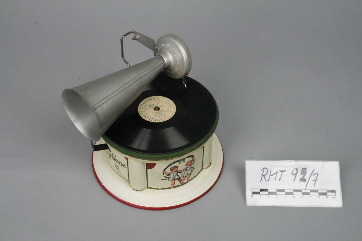 Leketøysgrammofon for avspilling av plater. Fjærmekanikk innvendig som trekkes opp for avspilling.
Nål og trakt som høytaler. 
Alle deler er i metall. Topplaten er malt grønn og limt rett på metall. Muligens en slags vinylmaling.