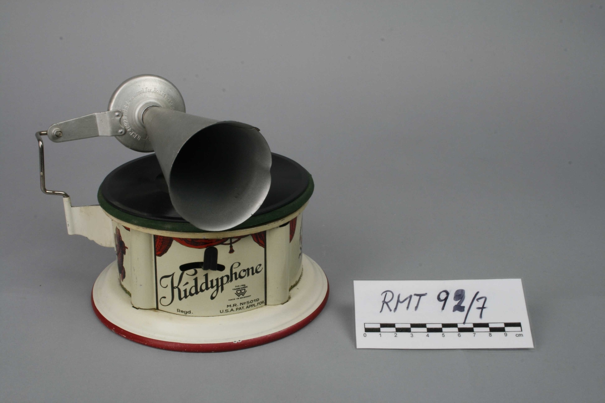 Leketøysgrammofon for avspilling av plater. Fjærmekanikk innvendig som trekkes opp for avspilling.
Nål og trakt som høytaler. 
Alle deler er i metall. Topplaten er malt grønn og limt rett på metall. Muligens en slags vinylmaling.