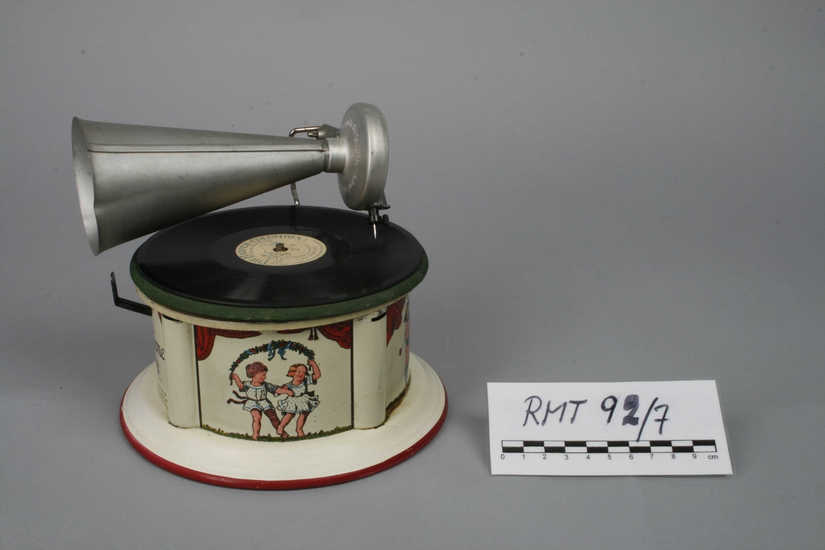 Leketøysgrammofon for avspilling av plater. Fjærmekanikk innvendig som trekkes opp for avspilling.
Nål og trakt som høytaler. 
Alle deler er i metall. Topplaten er malt grønn og limt rett på metall. Muligens en slags vinylmaling.