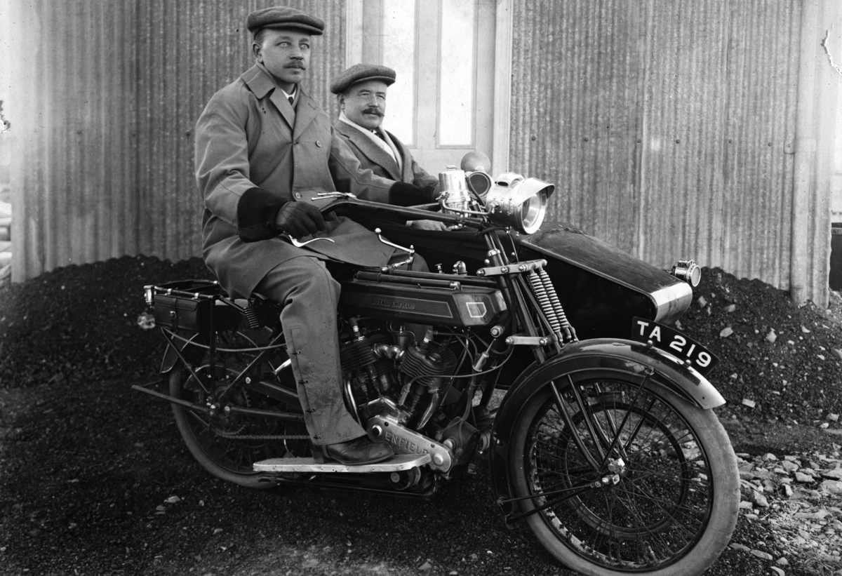 Dette er en Royal Einfield med sidevogn fra begynnelsen på 1920 tallet, (ant. 1922 modell). Skiltet tyder på at bildet er tatt i England da dette er et engelsk skilt fra samme tidsrom. Reg. nr. TA 219 Bernhard Skinderhaugen på sykkelen og Nils Skinderhaugen(?) i sidevogna. Bernhard Skinderhaugen emigrerte til Chicago i USA og ble en anerkjent fotograf. 