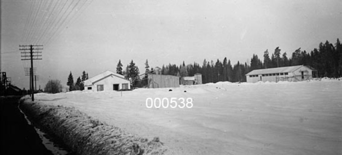 Fra Utne sag, høvleri og trelastforretning i Rygge i Østfold.  Fotografiet viser nyanlegget, som ble reist etter at det opprinnelige bruket i 1952 ble ekspropriert som en del av grunnen til Rygge flystasjon.  Opptaket er gjort vinterstid, fra en bar grusveg (til venstre i bildet), men mot et snødekt landskap der det ligger en del bygninger - blant annet saghus - på ei slette med skog i bakgrunnen. 