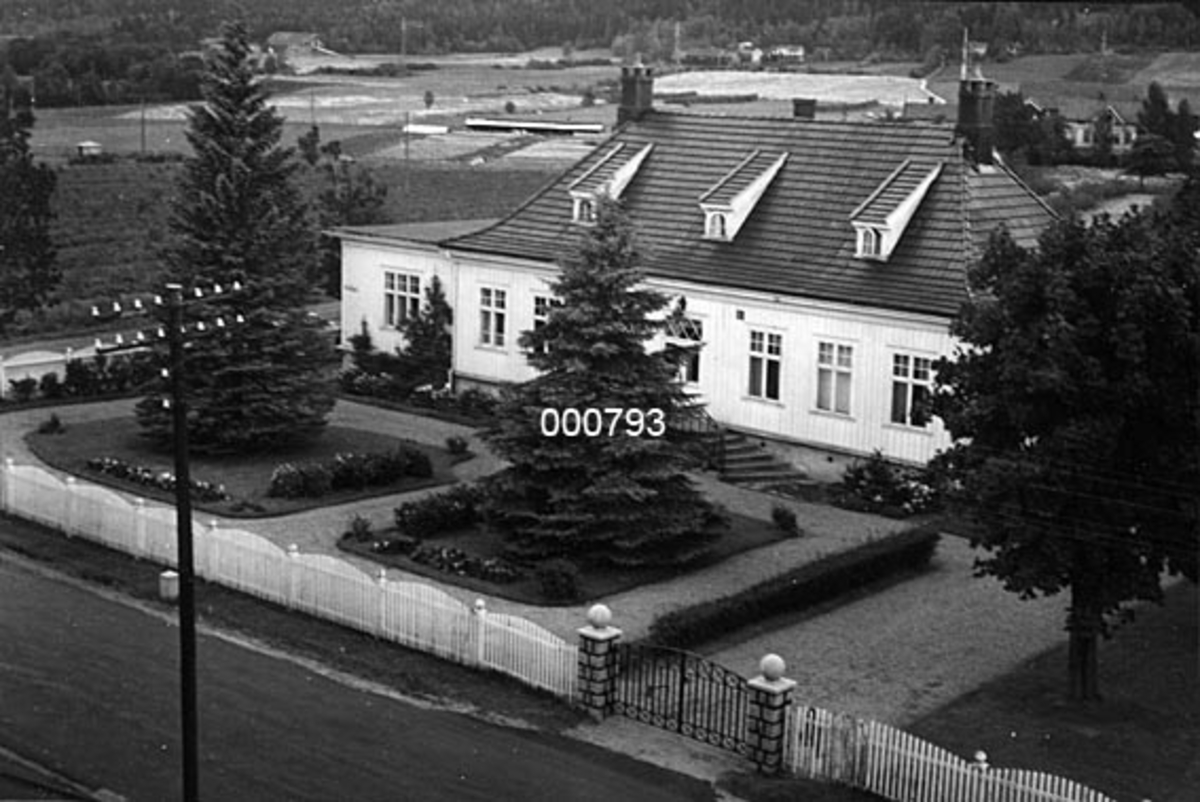 Eksteriør fra Bergskaug, et gardsbruk der bygningene i tunet ble brukt av tilstøtende A/S Buskerud Papirfabrikk på Åssida i Drammen (den gang i Lier kommune).  Fotografiet er antakelig tatt fra taket på fabrikken, over vegen mellom Drammen og Krokstadelva, mot våningshuset på Bergskaug.  Bygningen er oppført som et herregårdspreget enetasjes hus med panelte, kvitmalte yttervegger, jugendvinduer og med svaiet valmtak med arker.  Huset har to skorsteinspiper.  Foran en hage, med to rabatter med hvert sitt tre omgitt av blomster, og et stakittgjerde med smijernsport. 