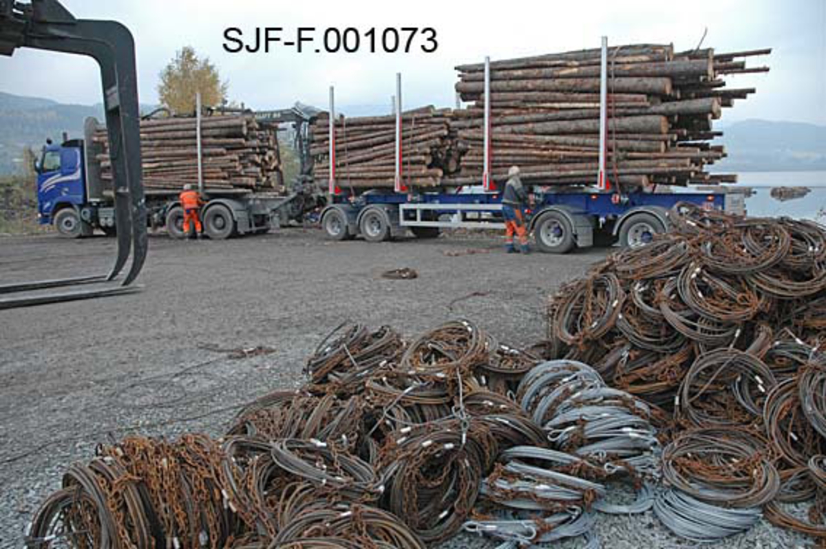 Fra Skiensvassdragets fellesfløtningsforenings utislagsplass på Nesøya ved Notodden.  Sentralt i bildet står en tømmerbil, som er kommet for å levere ubarket grantømmer til papirproduksjonen ved Union-fabrikken i Skien.  Sjåføren (til venstre) og en av Skiensvassdragets fellesfløtingsforenings medarbeidere (Tom Kaafjeld - til høyre) arbeider med bindsler ved lassene.  I forgrunnen ligger en haug av mer eller mindre rustne kabler ("bilband"), som knyttes rundt lassbuntene før de ble slått uti og skulle fløtes.  Til venstre i bildet ses gripeklørne på den 32 tonns gaffeltrucken som ble brukt til å løfte lassene av lastebilene. 