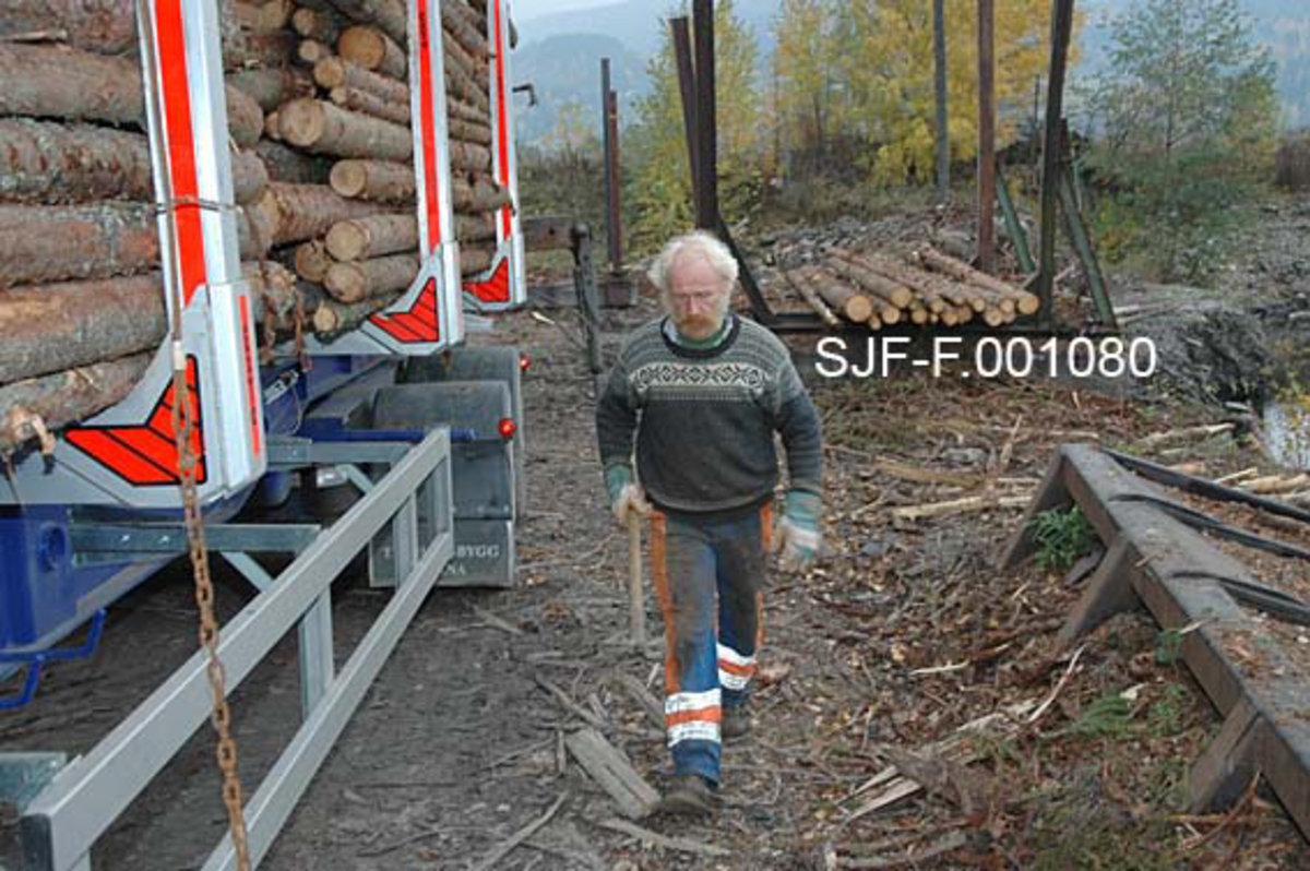 Fra Skiensvassdragets fellesfløtningsforenings utislagsplass på Nesøya ved Notodden.  Fotografiet viser Tom Kaafjeld, en av medarbeiderne i Skiensvassdragets fellesfløtningsforening, som nettopp har kastet et "bilbind" over et lastebillass med tømmer (jfr. SJF-F. 001078 og SJF-F. 001079).  Her er Kaafjeld på veg rundt lasset med en liten hake, som han skal bruke til å tre enden av bindet under lasset (jfr. påfølgende bildenummere).  Et bilbind besto av cirka ti meter vaier og cirka to meter med kjetting.  I enden av kjettingen var det et pæreformet lås.  Som en sikkerhetsforanstaltning ble låsene surret med tynn stålståd, såkalt blomstertråd.  Bilbindet skulle følge tømmeret fra utislaget til det var fløtet fram til papirfabrikken.  

Kaafjeld er iført svart strikkegenser med kvitt mønster og oransje og blå arbeidsbukser.  Han beskytter hendene ved å bruke arbeidshansker. 