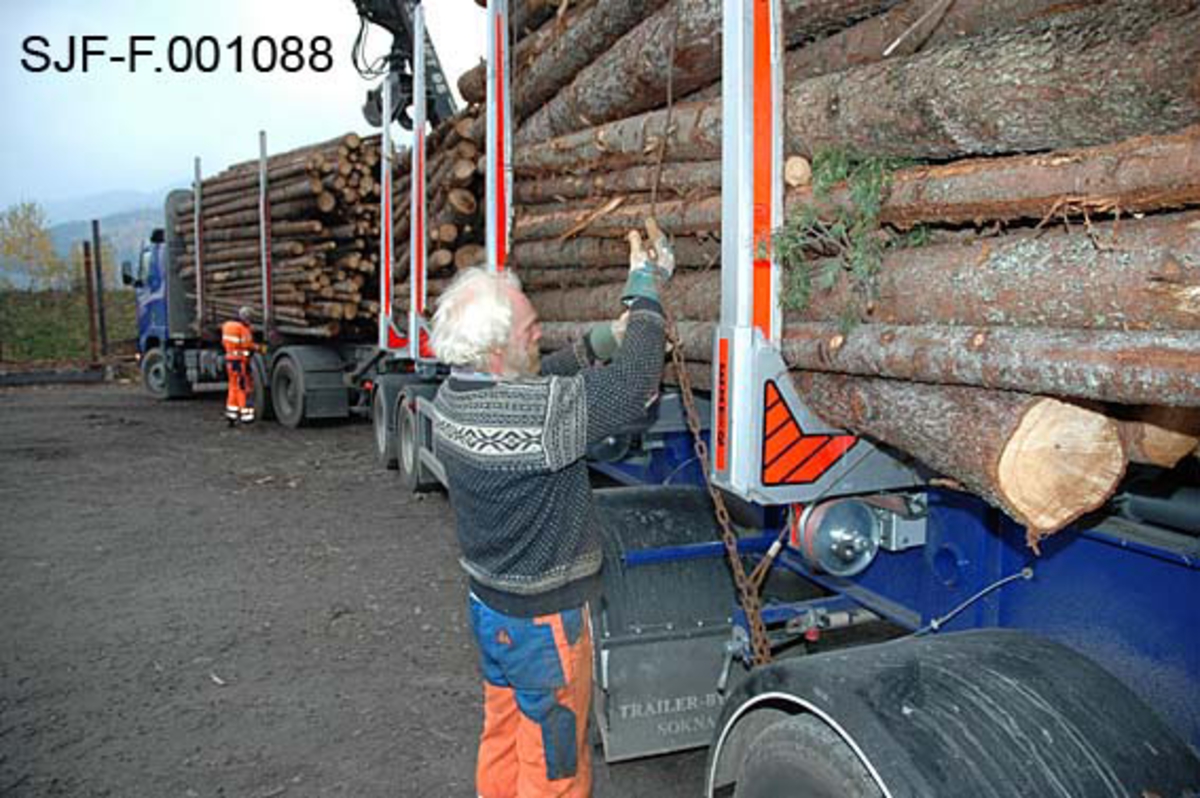 Fra Skiensvassdragets fellesfløtningsforenings utislagsplass på Nesøya ved Notodden.  Fotografiet viser Tom Kaafjeld, en av medarbeiderne i Skiensvassdragets fellesfløtningsforening, idet han låser et "bilbind" rundt et lastebillass med tømmer.  Et bilbind besto av cirka ti meter vaier og cirka to meter med kjetting.  I enden av kjettingen var det et pæreformet lås.  Som en sikkerhetsforanstaltning ble låsene surret med tynn stålståd, såkalt blomstertråd.  Bilbindet skulle følge tømmeret fra utislaget til det var fløtet fram til papirfabrikken.  

Kaafjeld er iført svart strikkegenser med kvitt mønster og oransje og blå arbeidsbukser.  Han beskytter hendene ved å bruke arbeidshansker. 