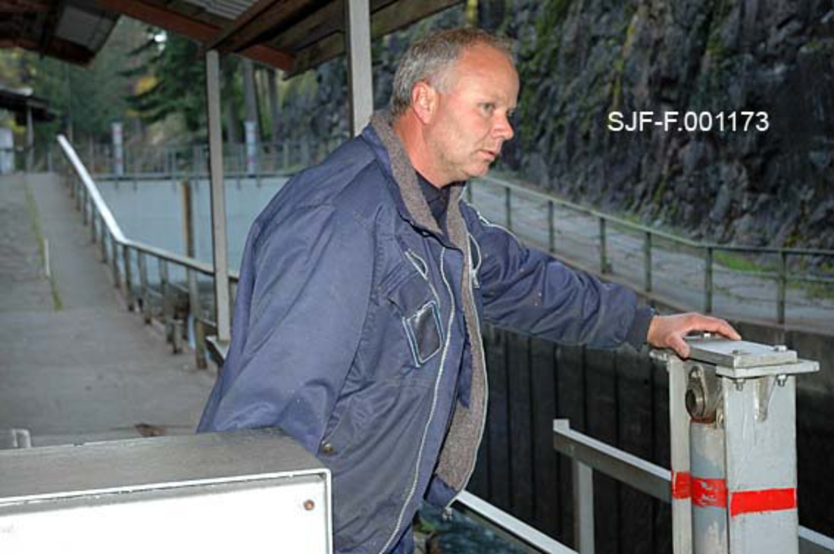 Geir John Kaasa ved betjeningspanel for sluseportene ved Løveid sluser i Skiensvassdraget.  Kaasa var ansatt i Telemarkskanalen.  Sammen med en kollega og to medarbeidere fra Skiensvassdragets fellesfløtningsforening sørget han for at tømmerbuntene kom vel gjennom slusene her.  Kaasa er kledd i blått arbeidstøy.  Han står med blikket vendt mot et av slusekamrene. 