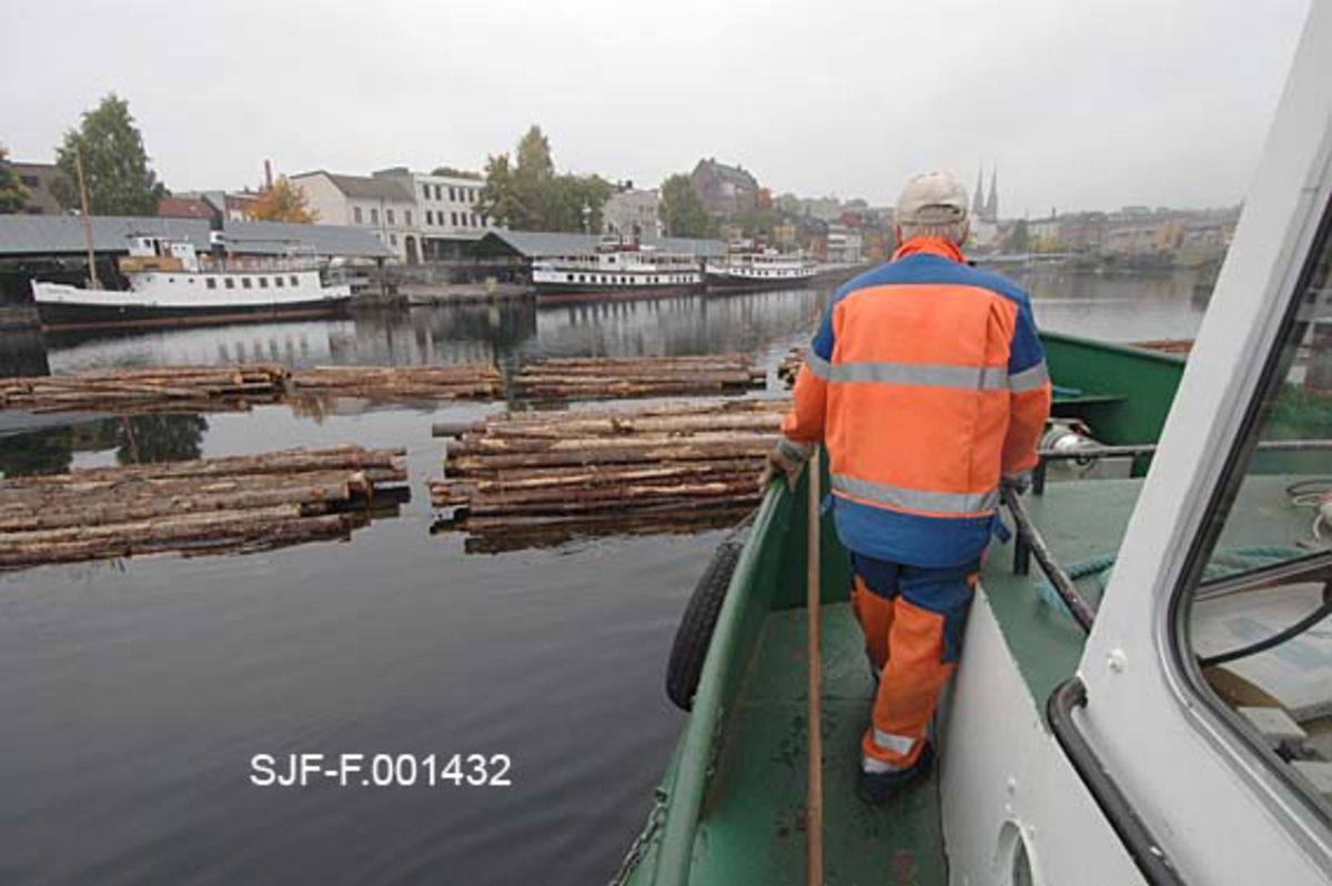 Torgrim Mork på dekket av slepebåten Triset.  Vi ser noe av styrhuset.  I bakgrunnen ligger et tømmerslep på vannet, og ved brygga på motastt side ligger tre veteanbåter.  Fotografiet skulle forakres ved land på Hjellevannet, ved den såkalte Damfossen, i påvente av at en kollega fra Skiensvassdragets fellesfløtingsforening med en mindre båt kom for å hente kortere lenker med tømmer for slusing gjennom Skien sluse til det nedenforliggende Bryggevannet og papirfabrikken Union. 