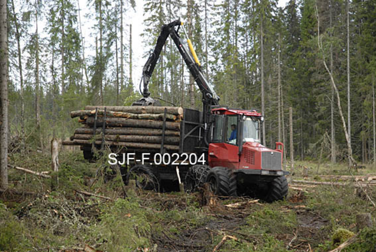 Ola Engens fire år gamle rammestyrte lassbærer, en Valmet-modell, i virksomhet i Ulvåsen i østre Elverum, 5. august 2009.  Fotografiet er tatt skrått forfra.  Maskinen har to akslinger på forparten, som bærer førerhuset, og to på bakparten, som bærer lasset.  På samtlige akslinger er det brede hjul med samme mønstring som bakhjulene på landbrukstraktorer.  Da dette fotografiet ble tatt var maskinen fullastet, og kom kjørende i oppkjørte spor på forholdsvis tørr mark. 