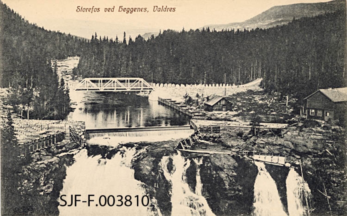 Storefoss ved Heggenes i Østre Slidre i Valdres.  Fotografiet er tatt i motstrøms retning, mot fossen, hvor vi ser den øvre delen av det kvitskummende fallet.  I overkant av det naturlige fallet later det til å være bygd en damterskel, og på begge sider aner vi kraftige, gråsteinfylte tømmerkistekonstruksjoner.  En slik skjerm – skådam – er det også langs østre elvebredd (til høyre) ovenfor dammen.  Innenfor skådammen ligger det ei laftet koie, og helt inne ved høyre bildekant ser vi en toetasjes bygningskonstruksjon med laftet førsteetasje og andreetasje i bordkledd bindingsverk.  Bygningens ytre gir ingen klare indikasjoner om hvilke funksjoner den rommet.  Til høyre i forgrunnen skimter vi litt av ei vannrenne med trevegger.  Ettersom denne renna hadde inntak nedenfor den nevnte løftedammen er det mer nærliggende å anta at det kan ha vært ei vassrenne til et nedenforliggende sag- eller møllebruk enn ei tømmerrenna som skulle lede fløtingsvirket uskadd forbi fossefallet.  Dette behøver imidlertid ikke å bety at slik renne ikke fantes.  Om lag 40-50 meter ovenfor dammen på fossebrinken krysser ei vegbru av støpejern elveløpet.  På østsida ser vi hvordan vegtraseen er oppbygd for å gi brua romslig høyde over vannspeilet.  Ettersom denne fyllinga har bratte sideflater, er vegbanen omkranset av stabbesteiner.  Landskapet ovenfor brua og vegtraseen preges av tett granskog.  I bakgrunnen skimter vi skogløse fjellrygger.  Storefoss ligger i Slidreåne, i det korte partiet som skiller den ovenforliggende Heggefjorden fra den nedenforliggende Volbufjorden. 