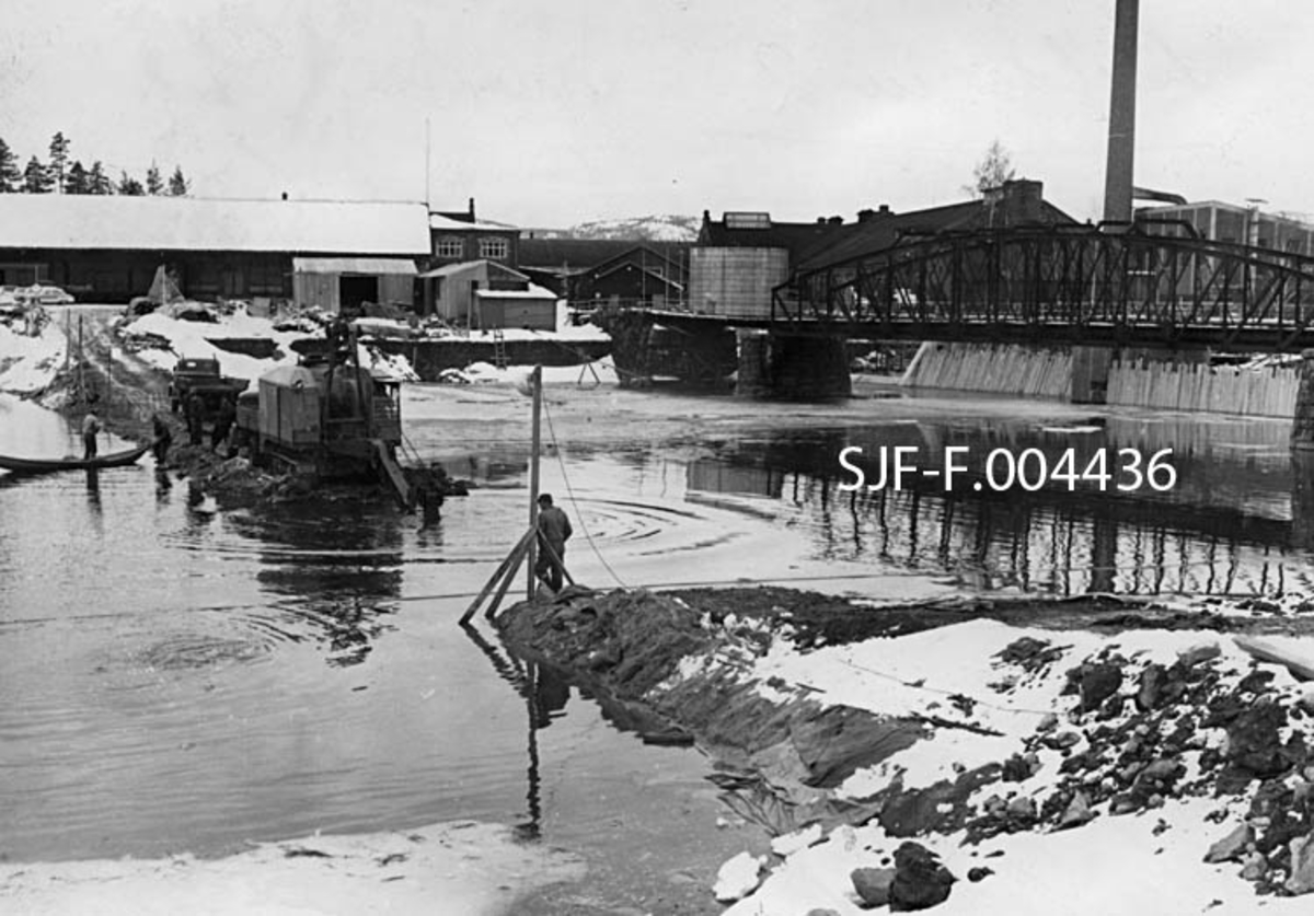 Fjerning av fangdam ovenfor Geithusfossen på Modum i Buskerud våren 1962.  Denne dammen ble bygd i forbindelse med at det foregående vinter ble arbeidet med en ny betongdam i sjølve Geithusfossen.  Her ser vi hvordan massen i fangdammen blir gravd opp med maskin og lesset opp på en ventende lastebil.  Bak gravemaskinen skimtes to slike biler.  En mann sto i en robåt på motstrøms side av fangdammen.  En annen mann fulgte arbeidet fra en posisjon ved en lyktestolpe på den delen av fangdammen som gjensto på vestre elvebredd.  Til høyre i bildet ser vi jernbanebrua på sidesporet fra Geithus stasjon på Randsfjordbanen til Drammenselvens Papirfabrikk.  Bakenfor skimter vi en del av bygningene ved bedriften, med lagerhall for avispapir og greaseproof (matpapir) til venstre, og med produksjonsbygninger til høyre. 