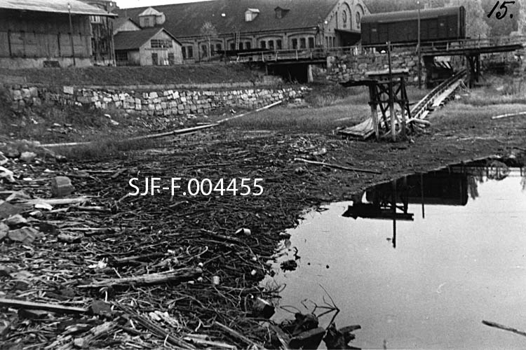 Strandareal ved Drammenselva, like ovenfor Geithusfossen på Modum i Buskerud.  Fotografiet er tatt ved ekstremt lav vannstand.  I øvre del av fotografiet ser vi en del av bygningene ved Drammenselvens Papirfabriker.  En skinnegang øverst til høyre i bildet er kjerraten som hadde blitt brukt til å trekke tømmer opp til fabrikken.  Kjerraten går under jernbanesporet, der det sto ei godsvogn.  Med slike vogner hentet man papirruller og pakker med greaseproof (matpapir), som ble transportert med tog til Geithus stasjon og derfra videre på Randsfjordbanen.  Strandområdet der dette fotografiet er tatt forandret karakter etter at det i 1960-61 ble sprengt en driftstunnel for et nedenforliggende kraftverk (Geithusfoss), og da det påfølgende år (1961-62) ble støpt en løftedam på toppen av fossen like nedenfor. 