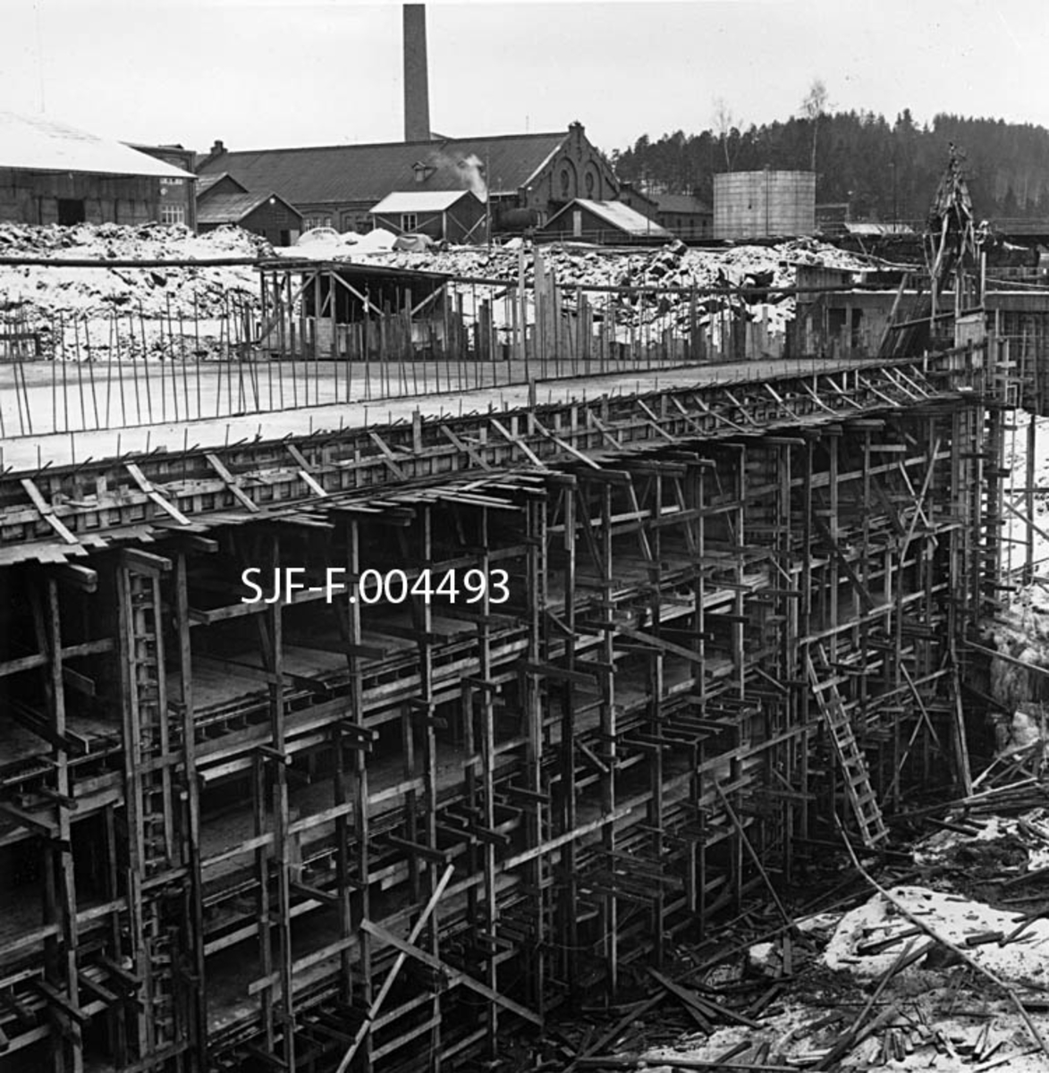 Støpearbeid ved inntaket til Geithusfoss kraftverk i byggeperioden, vinteren 1961.  Vannet til kraftproduksjonen ble hentet fra et punkt på Drammenselvas østre bredd ovenfor Geithusfossen.  Her ble det sprengt et traktformet dagbrudd, deretter en tunnel med tverrsnitt på om lag 70 kvadratmeter nedover mot fordelingsbassenget og kraftstasjonen drøyt 100 meter lengre nede.  Dette fotografiet viser hvordan det traktformete innløpspartiet ble overbygd med et dekke av armert betong og med et frontparti hvor det var rektangulære åpninger hvor vannet kunne trenge inn.  På denne fronten mot elva ble det seinere montert perforerte varegrinder av stål som skulle holde tømmer og andre objekter som kom flytende på elva unna driftsvannet til kraftverket.  Langs ytterkanten av det støpte dekket ble et lagt en skinnegang for ei kran som ble brukt til å renske grindene.  Så langt var imidlertid ikke arbeidet kommet da dette fotografiet ble tatt.  I bakgrunnen ser vi noe av bebyggelsen ved Drammenselvens Papirfabrikker, som hadde fallrettighetene i Geithusfossen.  Det var morselskapet Union Co. som sto for utbygginga, men kraftverket er seinere overtatt av Energiselskapet Buskerud Kraftproduksjon A/S. 