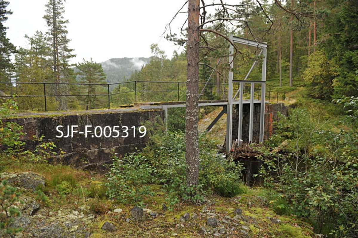 Vigdesjådammen ved utløpet av tjernet Vigdesjå i Kviteseid i Vest-Telemark.  Dammen er bygd av hoggen naturstein med krone av betong.  Dambrua har jernrekkverk på medstrøms side og ei stålramme for damlukene på motstrøms side.  Her ved utløpet av Vigdesjå renner Bygdaråi øst- og etter hvert nordøstover i bergrikt lende med Damåsan på nordsida og Kolakosi på sørsida, mot Høgfossen og Kolbjørnsrudfossen,  Fotografiet viser dammen omgitt av et landskap med blandingsskog (mye furu) og bergflater med lav- og lyngvegetasjon.  Bildet er tatt i sørøstlig retning mot damkonstruksjonen på motstrøms side. 