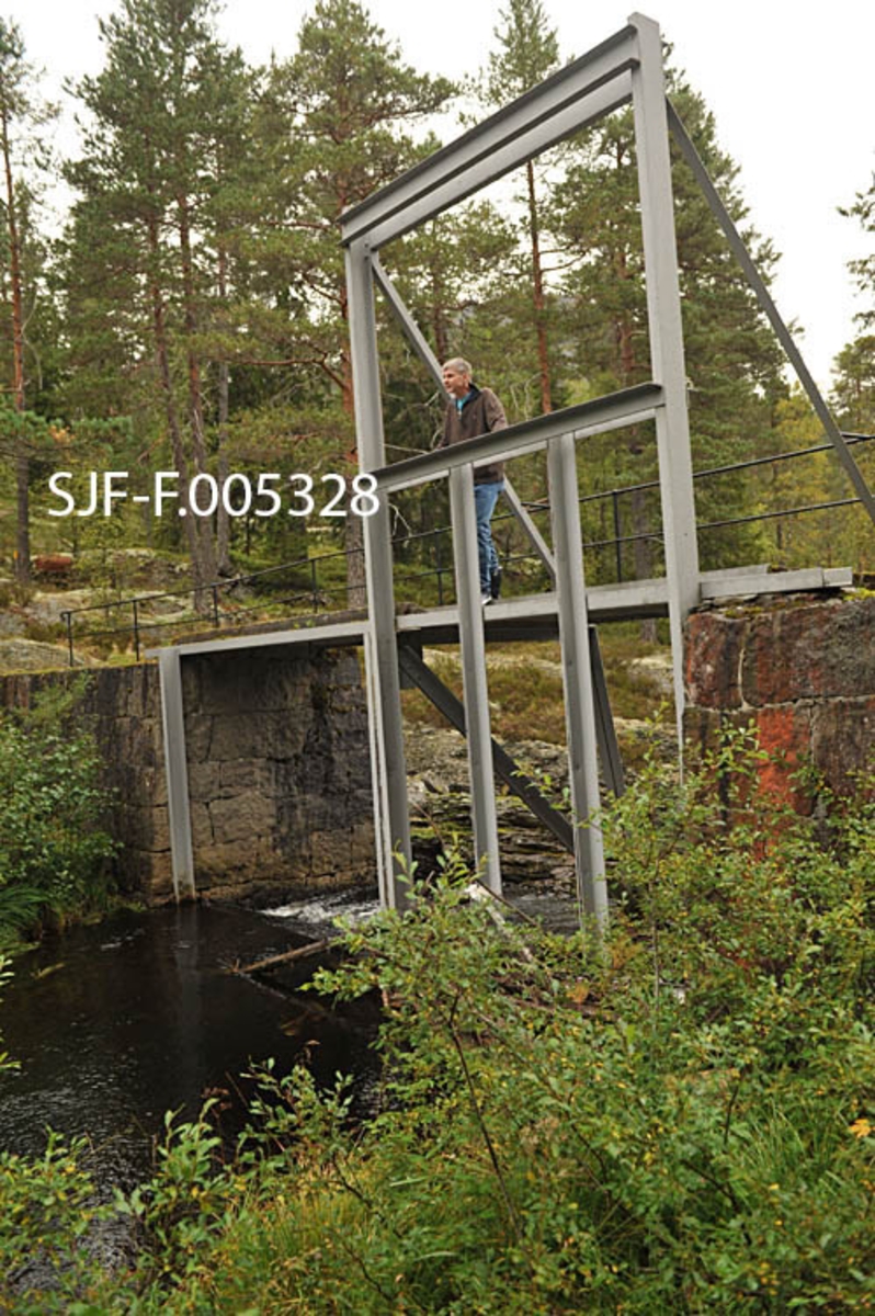 Vigdesjådammen ved utløpet av tjernet Vigdesjå i Kviteseid i Vest-Telemark.  Dammen er bygd av hoggen naturstein med krone av betong.  Dambrua har jernrekkverk på medstrøms side og ei stålramme for damlukene på motstrøms side.  Her ved utløpet av Vigdesjå renner Bygdaråi øst- og etter hvert nordøstover i bergrikt lende med Damåsan på nordsida og Kolakosi på sørsida, mot Høgfossen og Kolbjørnsrudfossen,  Fotografiet viser dammen omgitt av et landskap med blandingsskog (mye furu) og bergflater med lav- og lyngvegetasjon.  Bildet er tatt i nordlig retning, mot damkonstruksjonen og damåpningent Her var alle stengsler av tre borte på opptakstidspunktet, 40 år etter siste fløtingssesong.  Konservator Bjørn Bækkelund fra Norsk Skogmuseum sto på dambrua da bildet ble tatt. 