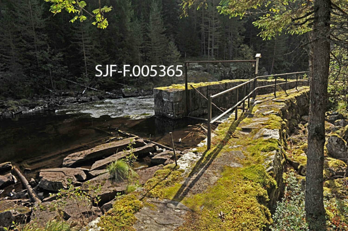 Innløpsrenna til tømmertunnelen ved Raufossen i Raua, elvestrekningen mellom Reisjåvatnet og Tinnsjøen i Øst-Telemark.  Fotografiet er tatt i medstrøms retning fra en posisjon like ovenfor innløpet innløpsrenna til den om lag 60 meter lange tunnelen, med innløpsrenna til høyre og den øvre delen av fosseløpet til venstre.  Som fotografiet viser var disse to løpene skilt av en kraftig, sementfuget gråsteinsmur.  Hensikten var dels å lede tømmeret mot tunnelen i stedet for mot fossen, hvor stokkene lett kunne splintres, dels å kunne begrense tilsiget av vatn til renna i flomperioder.  Ovenfor innløpet til renna skal det ha vært lense, støttet av to kraftige, steinfylte tømmerkistekar.  Rekkverket på landsida (til høyre i bildet) ble oppsatt i forbindelse med at det ble opparbeidet kultursti her, altså lenge etter at fløtingsaktiviteten opphørte. 