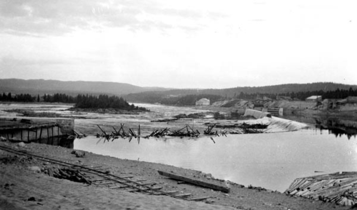 Skjefstadfossen i Glomma, nærmere bestemt i Heradsbygda i Elverum.  Fotografiet er tatt sommeren 1910, like etter at første fase i kraftutbygginga på dette stedet var avsluttet. Bildet er tatt fra elvas østre bredd, der det fortsatt lå en skinnegang som antakelig var brukt til å trille fram materialer i forbindelse med støpinga av den lange, buete dammen i den sentrale delen av bildeflata.  Denne dammen skulle fange opp vannmasser til Skjefstadfossen kraftverk, som hadde sitt vanninntak gjennom turbinrør ved den vestre elvebredden, og sine generatorer i en bygning vi ser drøyt lengre nede på vestsida av elva.  Dette fotografiet er antakelig tatt for Christiania Tømmerdirektion, som hadde behov for å dokumentere hvordan fløtingstømmeret hadde filtret seg sammen i vaser under den østre delen av damkonstruksjonen.  Tømmerhauger på et slikt sted innebar farlig arbeid for fløterne og ekstrakostnader for organisasjonen som var ansvarlig for at virket kom vel fram til den tømmerkjøpende industrien lengre nede i vassdraget. 

Litt mer informasjon om kraftanlegget ved Skjefstadfossen finnes under fanen «Andre opplysninger». 