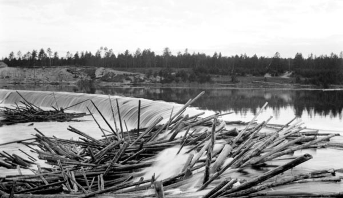 Tømmerfløting ved Skjefstadfossen i Glomma, antakelig sommeren 1910.  Fotografiet er tatt fra det høye og kraftige landkaret som forbandt den drøyt 300 meter lange, støpte damkonstruksjonen med vassdragets østre bredd.  Da dette fotografiet ble tatt rant det vann over både den østre delen av den buete damkrona, som var den laveste, og den midtre, som var en aning høyere.  På begge steder har det bygd seg opp hauger av fløtingsvirke, antakelig fordi tømmeret falt fra dammen og ned i et parti der elvebotnen var steinete og ujamn.  Lengst øst, der de største tømmermengdene gikk, har tømmerhaugen bygd seg opp i sjølve dammen.  Det å løsne slike tømmervaser, mens vannføringa ennå var høy og mulighetene for videre fløting var gode, var farefullt arbeid for involverte mannskaper og en kostnadsdrivende faktor for Kristiania Tømmerdirektion.  Følgelig er det grunn til å anta at fotografiet ble tatt for å dokumentere slike vanskeligheter overfor kraftutbyggerne, som hadde plikt til å holde andre aktører med hevd på bruk av vassdragets skadesløse. 