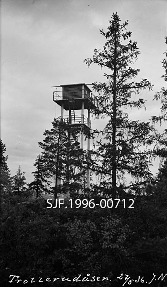 Skogbrannvaktstasjonen på Trollerudåsen i Spydeberg.  Brannvakttårnet på dette stedet ble reist i 1935, som en erstatning for eldre brannvaktposter på Kjerringa, Spydeberg varde og Bjørnåsen.  Trollerudås-tårnet er i hovedsak bygd av stål og betong, værbestandige materialer som skulle gi konstruksjonen lang levetid.  Tårnet besto av fire loddrette betongsøyler med tre horisontale avsatser mellom.  Samtlige avsatser hadde stålrekkverk. På den øverste avsatsen var det bygd ei lita hytte, en utkikkspost, av tre.  Atkomsten til hytta skjedde ved hjelp av stiger som var ført gjennom åpninger i de ulike avsatsdekkene.  Dette fotografiet ble tatt på avstand og viste hvordan tårnet var plassert på et berg der det vokste litt forkrøplet gran- og furuskog i revnene. 