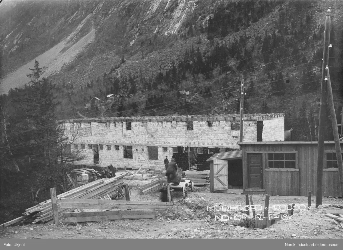 Murhusbebyggelsen på Krosso under oppføring. Byggets andre etasje er nesten ferdig reist. Hauger med materialer og murstein flere steder på byggeplassen. To byggearbeidere i arbeid. Bebyggelse på sørsiden av juvet i bakgrunnen.