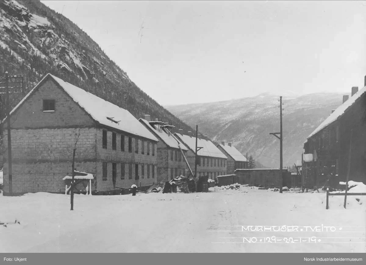 Bygging av fire murhus stående på rekke på Tveito på nordsiden av gaten Tveito Alle. Byggene på sørsiden av gaten er nær ferdigstilt. Lagerbygg stående midt i gaten mellom bygningene. Nyplantede trær langs gaten.