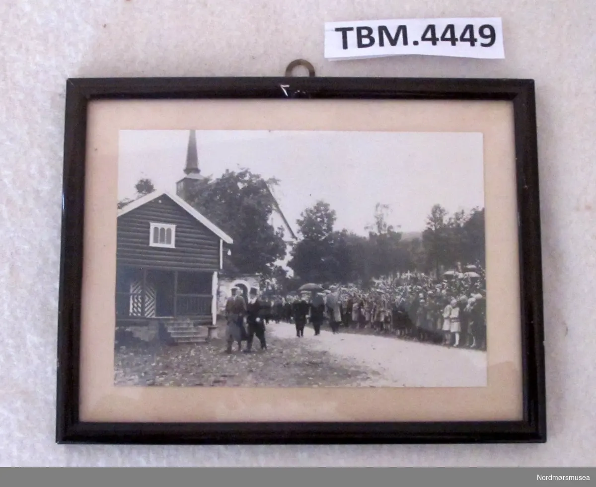 Fotografi i smal, svart ramme. Bildet viser kong Haaakon og Prost Sigurd Fjær på veg frå kyrkja til prestegarden. I bakgrunnen ei stor menneskemengde finkledde og med flagg.