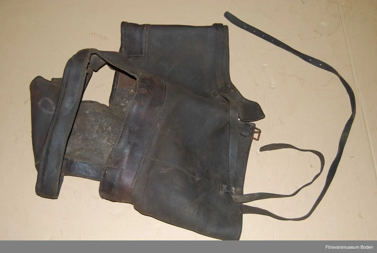 Kapell troligen för pjäs eller liknande. Av svart läder, med spännen. Närmast rund form med två rektangulära öppningar.