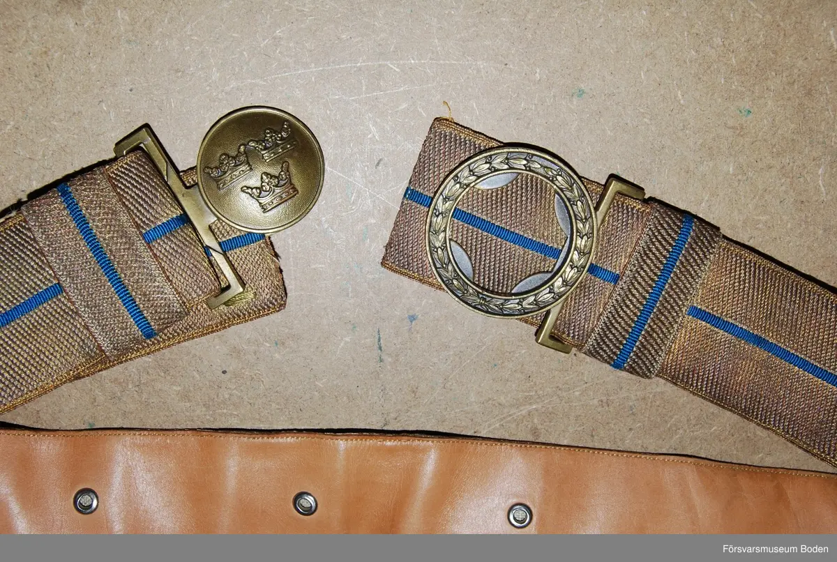 Bars till uniform m/1939 och m/1952 vid stor högtidsdräkt. Vävd av bronsfärgad tråd med smal blå rand inlagd. Spänne med tre kronor i brons, delat i två delar. För modellexemplar med utförlig beskrivning, se AM.004633.