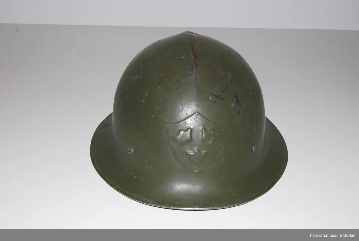 Olivgrön stålhjälm av typen med bredare och plattare brätte, den till vänster på bild 6 som jämför båda typerna av hjälm m/1921. Med insats bestående av tre läderkuddar som hålls ihop med skorem. Hakband av läder. Framtill tre kronor i relief, inga dekaler på sidorna. Upptill en förstärkningspressning i form av en kam.