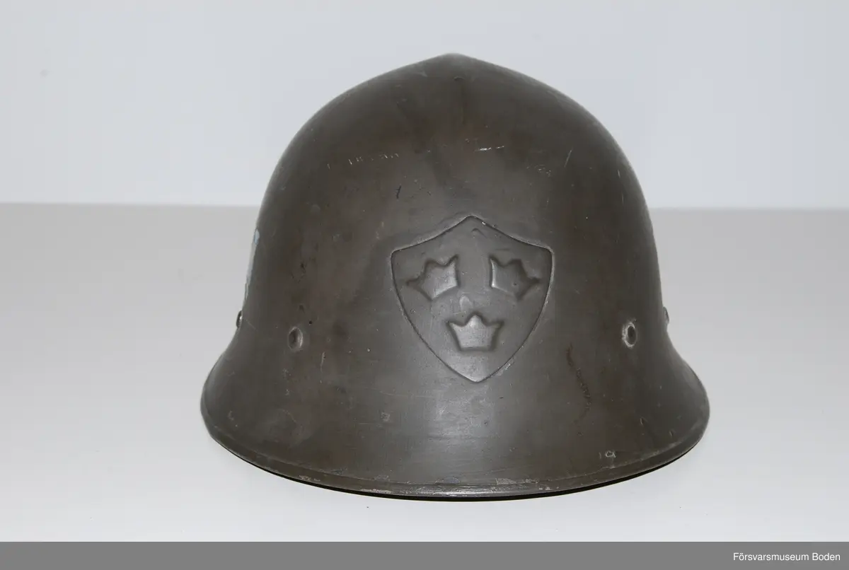 Mörkt grågrön stålhjälm av typen med smalare och högre brätte, den till höger på bild 6 som jämför båda typerna av hjälm m/1921. Med insats bestående av tre läderkuddar som hålls ihop med skorem. Hakband av läder. Framtill tre kronor i relief, tre-kronor dekaler på sidorna. Upptill en förstärkningspressning i form av en kam.