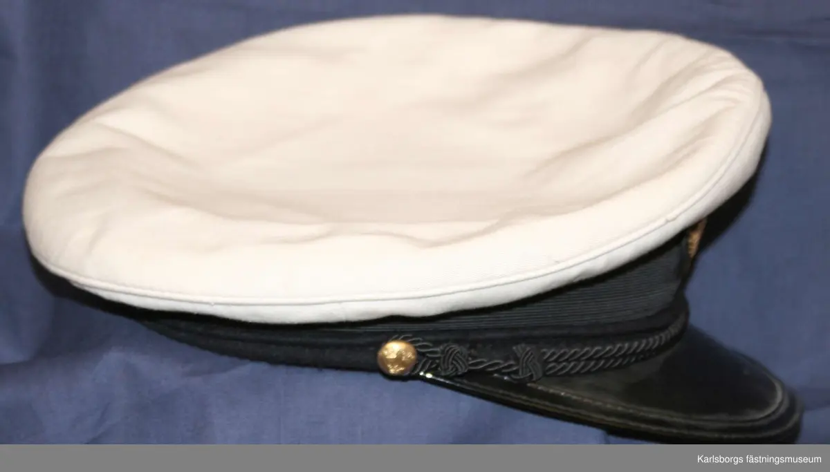 Storlek: 57
Skärmmössa m/1930 Ubef vit möskulle. Mössband av svart räfflat silke. Svart lackerad skärm. Flygemblem i guld- hakrem fäst vid 14 mm uniformsknappar.