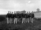 En grupp militärer med slutvärnet vid Karlsborgs fästning i bakgrunden, fotograferat i början på 1900-talet.