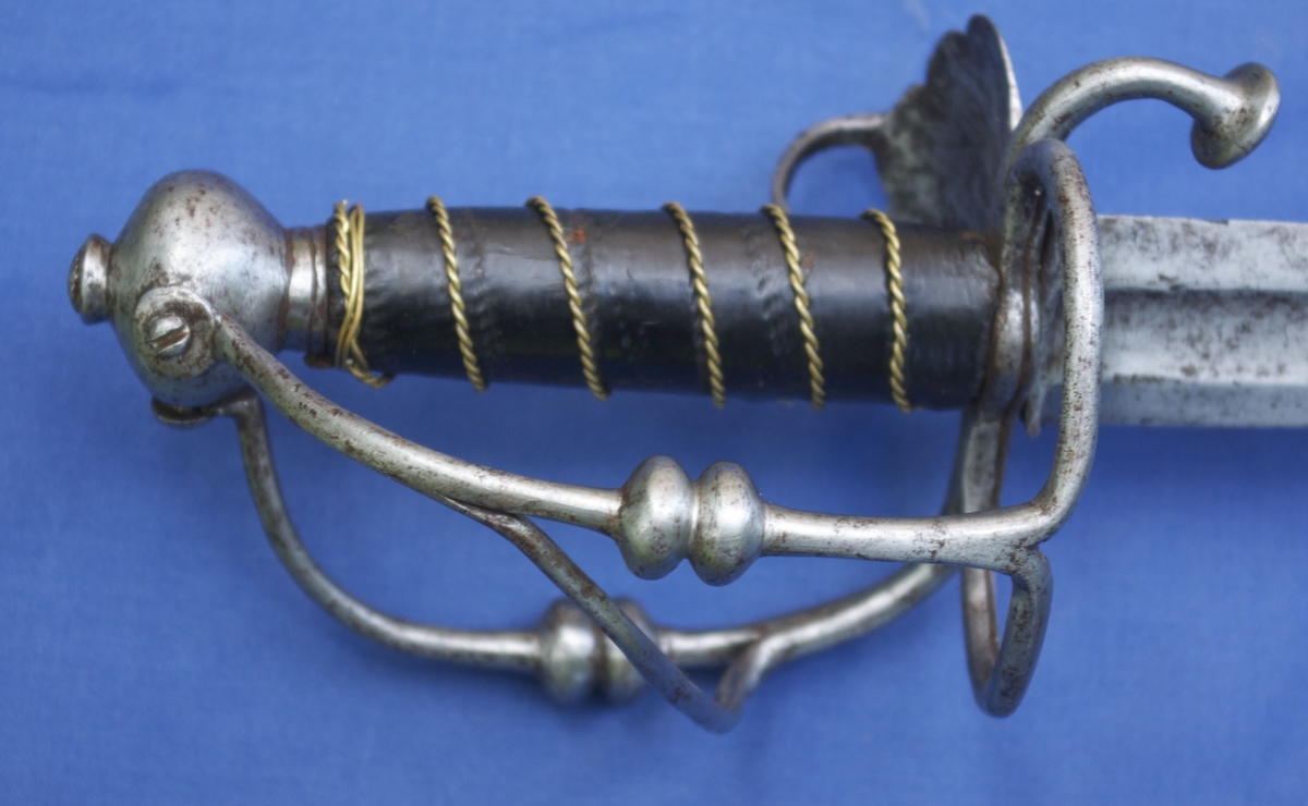 Tveegget sverd med hulslipt klinge og blodrand som tar til 9 cm fra spissen. Sverdgrepet har jernstenger til håndbeskyttelse. Sverdgrepets håndbeskyttelse er kunstferdig smidd av ett stykke. Parerstangen, som vel må være nedbøyd senere, ender i en knapp. Plate bare på tommelfingersiden, med fire furer som slutter i et innhakk i kanten, mellom furene gravert dekor av små ringer, platen har tommelfingerbøyle. På høyre side er i stedet for plate utsmidd en jernring som går over i en stang, den har to kuler midt på, danner en bøyle til beskyttelse for håndbaken og er skrudd fast på knappen. Den ordinære bøyle har også to kuler på midten, nedenfor kulen deler den seg og sender en buet stang over på håndbakbøylen, er skrudd fast til knappen. Rund knapp av jern, ender i en hals der tangen er klinket. Trekavle med brunt lær omviklet med snodd messingtråd i seks vindinger.
