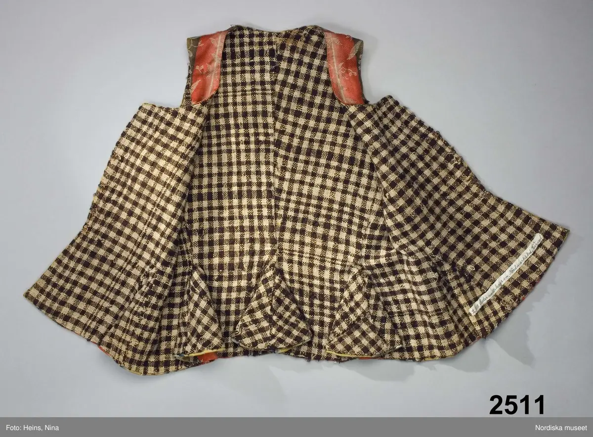 Livstycke med långt skört, av finaste kamgarnsylle i satin, randigt i varpled i breda bårder i tonande nyanser av rött, grönt, gult och blått med invävda blomsterslingor i vitt som ligger på en extra mönstervarp. Tygkvaliten kallas blommerad kalmink och har från början varit starkt glättat. 1700-talsmodell med 2 framstycken/sidstycken med skörtveck i den bakåtlagda sidsömmen och infälld kil i framstycket. Framkanterna något svängda. 2 ryggstycken med skörtveck i baksömmen. Dold knäppning fram med 4 par hakar och hyskor av järntråd. Foder av hemvävt rutigt linne i gles tuskaft, rutigt i mörkbrunt och ofärgat lingarn.
Anm. Något solkigt, på ena bakstycket en stoppning i midjelinjen. Nedersta haken saknas.
Kalmink blev modernt i slutet av 1700-talet till högtidsdräkter främst i bröllopsplagg. Det var ett köpetyg som kom från England där Norwich vävde de allra finaste kvaliteterna. Förmodligen inkommet via gränshandel med Norge.
/Berit Eldvik 2007-06-11