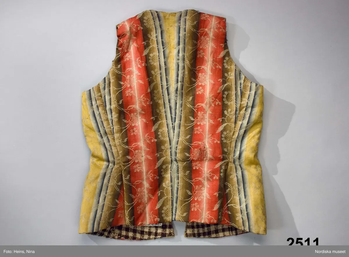 Livstycke med långt skört, av finaste kamgarnsylle i satin, randigt i varpled i breda bårder i tonande nyanser av rött, grönt, gult och blått med invävda blomsterslingor i vitt som ligger på en extra mönstervarp. Tygkvaliten kallas blommerad kalmink och har från början varit starkt glättat. 1700-talsmodell med 2 framstycken/sidstycken med skörtveck i den bakåtlagda sidsömmen och infälld kil i framstycket. Framkanterna något svängda. 2 ryggstycken med skörtveck i baksömmen. Dold knäppning fram med 4 par hakar och hyskor av järntråd. Foder av hemvävt rutigt linne i gles tuskaft, rutigt i mörkbrunt och ofärgat lingarn.
Anm. Något solkigt, på ena bakstycket en stoppning i midjelinjen. Nedersta haken saknas.
Kalmink blev modernt i slutet av 1700-talet till högtidsdräkter främst i bröllopsplagg. Det var ett köpetyg som kom från England där Norwich vävde de allra finaste kvaliteterna. Förmodligen inkommet via gränshandel med Norge.
/Berit Eldvik 2007-06-11