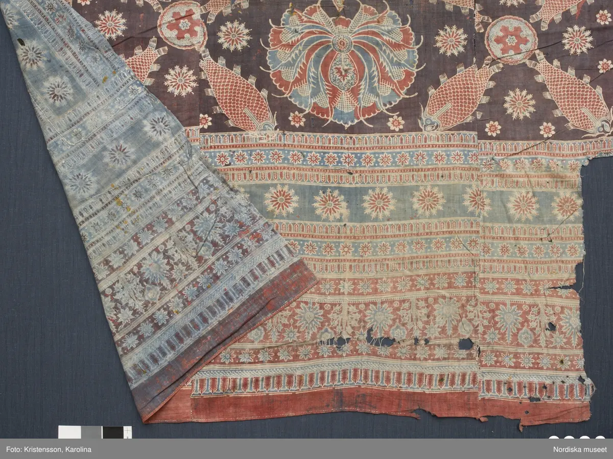 Stort tygstycke sammansytt av 3 våder bomullslärft  á 54 cm bredd. Hopsydda med vitt handspunnet lingarn. Tryckt mönster i rött och blått på vitt underlag.. I mitten stort cirkelmotiv fylld med mönster och i mitten en stjärna, åt fyra håll en triangel ut från cirkeln och mot 4 större snäckliknande ornament. I varje hörn motiv med 4 fiskar med stjärtarna in mot en cirkel, Kantbårder bestånde av olikmönstrade ränder i olika bredder.  Trycket är gjort med tryckstockar och smalare tryckstämplar i kantbårderna där tydliga blockskarvar syns.  Tekniken är den gamla indiska med betmålning av de partier som skall bli röda och därefter doppning i krappbad, tvättning av tyget före blåfärgning som görs så att vita och röda parteier täcks med en pasta före färgning i indigokyp. På baksidan är tyget mycket blåare än på framsidan. Spegelns botten har färgats först röd och därefter blå  så att den fått en mörklila , lätt flammig nyans. Tyget är skört och trasigt på flera ställen, längs hela högersidan är det söndigt vilket kan vara musätet. En del mindre hål har lappats med  gråblått siden på baksidan eller på ett ställe med en bit av själva tygtrycket. Även blekt.
Insamlat av Nils Månsson-Mandelgren vid hans resor i Västergötland, inga uppgifter finns.
 /Berit Eldvik 2013-06