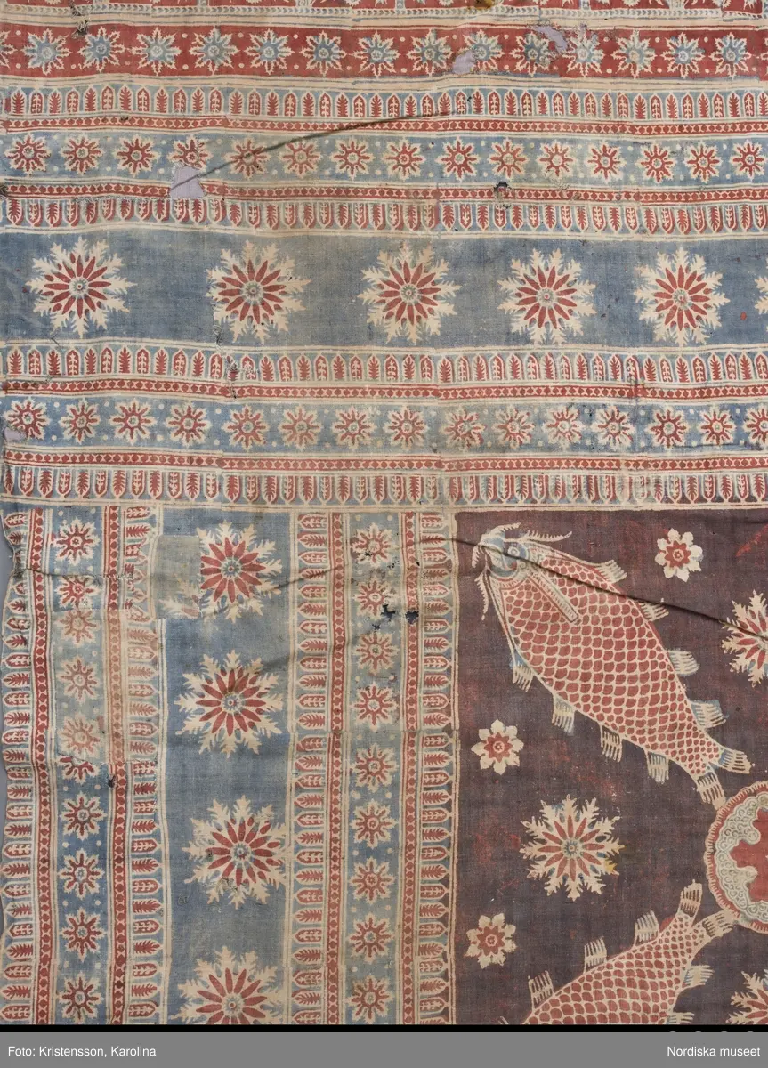 Stort tygstycke sammansytt av 3 våder bomullslärft  á 54 cm bredd. Hopsydda med vitt handspunnet lingarn. Tryckt mönster i rött och blått på vitt underlag.. I mitten stort cirkelmotiv fylld med mönster och i mitten en stjärna, åt fyra håll en triangel ut från cirkeln och mot 4 större snäckliknande ornament. I varje hörn motiv med 4 fiskar med stjärtarna in mot en cirkel, Kantbårder bestånde av olikmönstrade ränder i olika bredder.  Trycket är gjort med tryckstockar och smalare tryckstämplar i kantbårderna där tydliga blockskarvar syns.  Tekniken är den gamla indiska med betmålning av de partier som skall bli röda och därefter doppning i krappbad, tvättning av tyget före blåfärgning som görs så att vita och röda parteier täcks med en pasta före färgning i indigokyp. På baksidan är tyget mycket blåare än på framsidan. Spegelns botten har färgats först röd och därefter blå  så att den fått en mörklila , lätt flammig nyans. Tyget är skört och trasigt på flera ställen, längs hela högersidan är det söndigt vilket kan vara musätet. En del mindre hål har lappats med  gråblått siden på baksidan eller på ett ställe med en bit av själva tygtrycket. Även blekt.
Insamlat av Nils Månsson-Mandelgren vid hans resor i Västergötland, inga uppgifter finns.
 /Berit Eldvik 2013-06