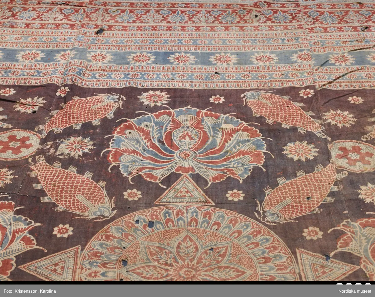 Stort tygstycke sammansytt av 3 våder bomullslärft  á 54 cm bredd. Hopsydda med vitt handspunnet lingarn. Tryckt mönster i rött och blått på vitt underlag.. I mitten stort cirkelmotiv fylld med mönster och i mitten en stjärna, åt fyra håll en triangel ut från cirkeln och mot 4 större snäckliknande ornament. I varje hörn motiv med 4 fiskar med stjärtarna in mot en cirkel, Kantbårder bestånde av olikmönstrade ränder i olika bredder.  Trycket är gjort med tryckstockar och smalare tryckstämplar i kantbårderna där tydliga blockskarvar syns.  Tekniken är den gamla indiska med betmålning av de partier som skall bli röda och därefter doppning i krappbad, tvättning av tyget före blåfärgning som görs så att vita och röda parteier täcks med en pasta före färgning i indigokyp. På baksidan är tyget mycket blåare än på framsidan. Spegelns botten har färgats först röd och därefter blå  så att den fått en mörklila , lätt flammig nyans. Tyget är skört och trasigt på flera ställen, längs hela högersidan är det söndigt vilket kan vara musätet. En del mindre hål har lappats med  gråblått siden på baksidan eller på ett ställe med en bit av själva tygtrycket. Även blekt.
Insamlat av Nils Månsson-Mandelgren vid hans resor i Västergötland, inga uppgifter finns.
 /Berit Eldvik 2013-06